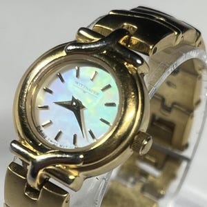 May include: A gold-coloured Wittnauer wristwatch with a mother-of-pearl face. The watch has a round dial with black markers and hands, and a gold-coloured strap. The watch is displayed on a clear stand.