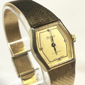 May include: A gold-toned Currier wristwatch with a hexagonal face and a textured band. The watch face displays the brand name, "Currier", and "17 Jewels". The band is a woven style. The watch is a vintage style.