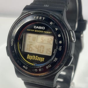 May include: Black Casio Depth Gauge watch with a digital display showing the time. The watch face has a black bezel with white and yellow markings, including depth measurements in feet. The watchband is black.