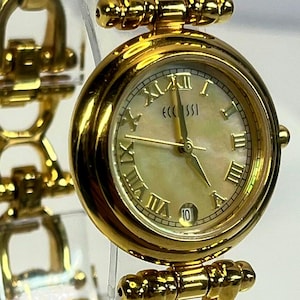 May include: A gold-toned wristwatch with a round face and Roman numeral hour markers. The watch features a mother-of-pearl dial and a date display at the 10 o'clock position. The watch band is also gold-toned and has a decorative design.
