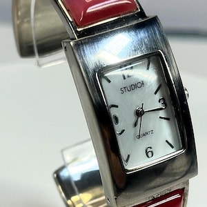 May include: A silver-toned Studio Quartz watch with a white face and black hands. The watch band features green, blue, and red rectangular accents. The watch has a rectangular face with the numbers 12, 3, 6, and 9.