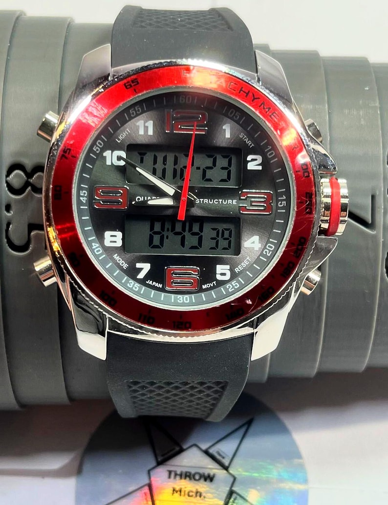 Peut inclure: Montre num&eacute;rique noire et argent&eacute;e avec une lunette rouge. Le cadran affiche l'heure et comporte plusieurs boutons. Le mot "CHYME" est imprim&eacute; sur le cadran. Le bracelet est en caoutchouc noir.