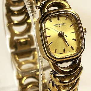 May include: Gold-toned Wittnauer watch with a square face and matching linked band. The watch face displays the Wittnauer brand name and has black hour markers. The band is composed of curved, interlocking links. The watch is displayed on a clear, curved surface.