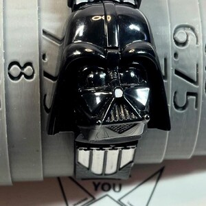 Pu&ograve; includere: Un orologio nero di Darth Vader con cinturino nero abbinato. Il quadrante dell'orologio &egrave; una replica dettagliata del casco di Darth Vader. L'orologio &egrave; posto su uno sfondo grigio con numeri.