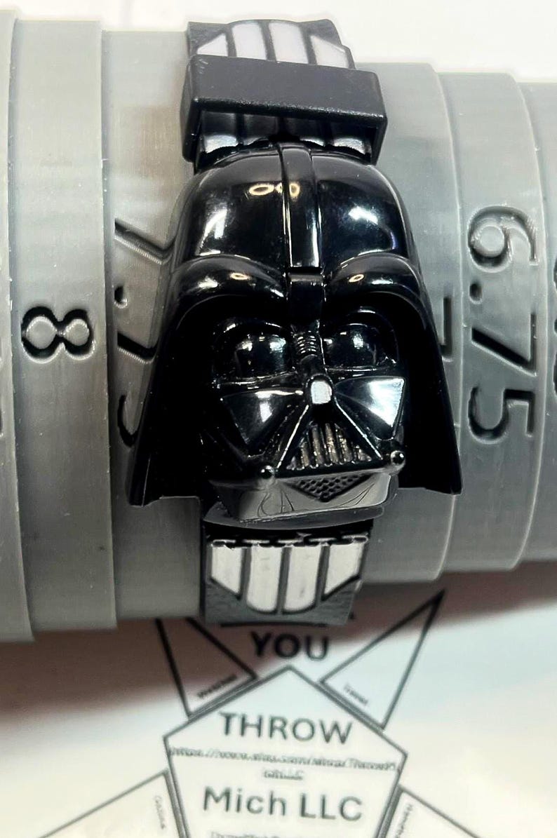 Pu&ograve; includere: Un orologio nero di Darth Vader con cinturino nero abbinato. Il quadrante dell'orologio &egrave; una replica dettagliata del casco di Darth Vader. L'orologio &egrave; posto su uno sfondo grigio con numeri.