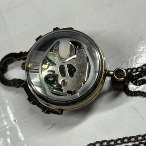May include: A steampunk-style pendant necklace featuring a clear, spherical watch face revealing the internal mechanism. The watch is encased in a bronze-colored metal frame and attached to a chain. The design includes decorative clasps and a dark chain.