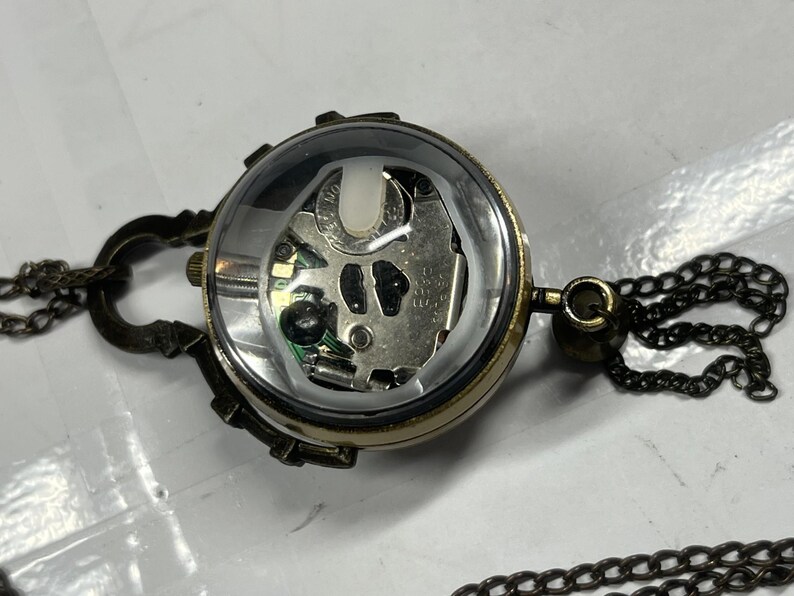 May include: A steampunk-style pendant necklace featuring a clear, spherical watch face revealing the internal mechanism. The watch is encased in a bronze-colored metal frame and attached to a chain. The design includes decorative clasps and a dark chain.