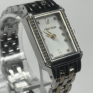 May include: A rectangular Armitron watch with a silver and gold band. The watch face is white with gold hands and small diamond accents. The bezel is silver with a row of small crystals.