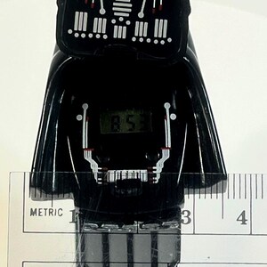 Pu&ograve; includere: Un orologio digitale nero a forma di casco di Darth Vader di Star Wars. Il quadrante mostra l'ora. L'orologio &egrave; misurato con un righello, mostrando una lunghezza di circa 10 cm.