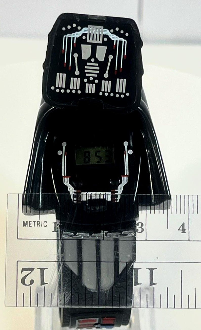 Pu&ograve; includere: Un orologio digitale nero a forma di casco di Darth Vader di Star Wars. Il quadrante mostra l'ora. L'orologio &egrave; misurato con un righello, mostrando una lunghezza di circa 10 cm.