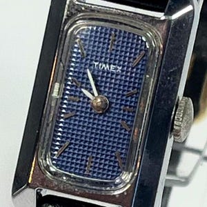May include: A vintage Timex watch with a rectangular silver-tone case and a navy blue patterned dial. The watch features silver hands and a black leather strap. The word "TIMEX" is printed on the dial.