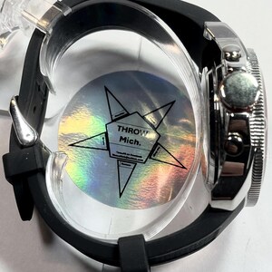 Peut inclure: Gros plan d'une montre argent&eacute;e avec un bracelet en caoutchouc noir. Le cadran de la montre est partiellement visible, et la lunette a un design textur&eacute;. La montre est maintenue dans un support en plastique transparent, avec le texte "THROW Mich."