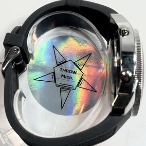 Peut inclure: Une montre-bracelet noire avec une lunette argent&eacute;e et un bracelet en caoutchouc noir. Le cadran de la montre est partiellement visible, et le dos de la montre a un autocollant holographique avec le texte "THROW Mich."
