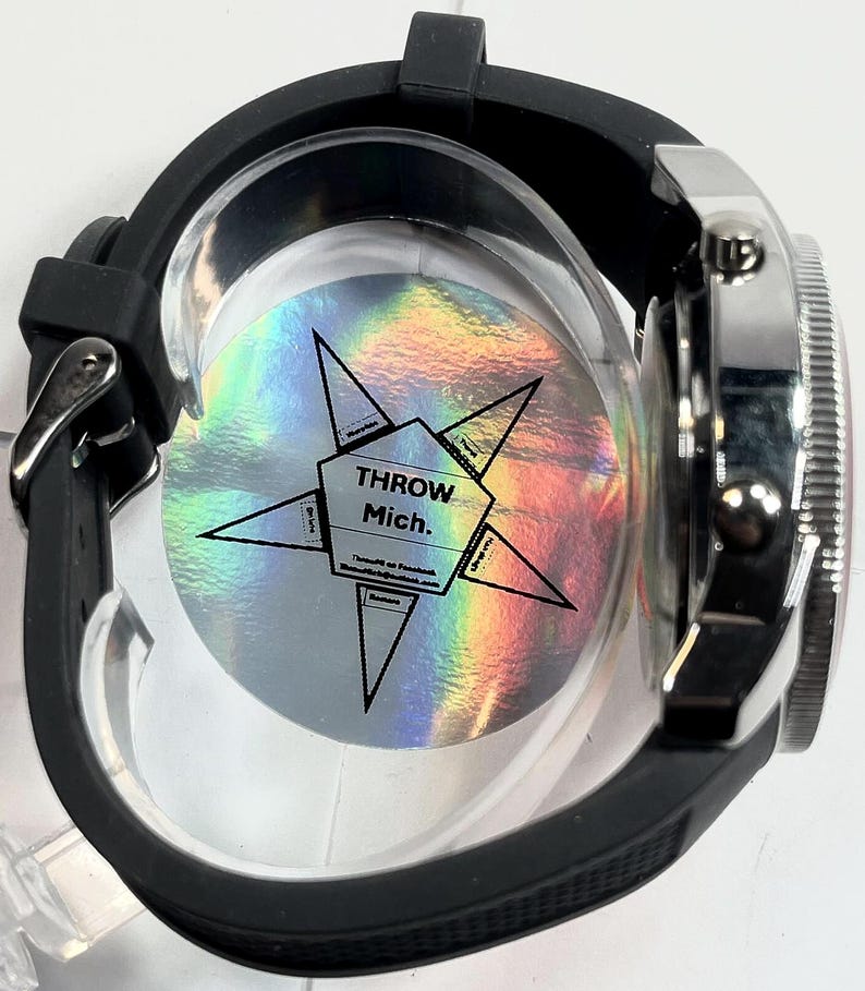 Peut inclure: Une montre-bracelet noire avec une lunette argent&eacute;e et un bracelet en caoutchouc noir. Le cadran de la montre est partiellement visible, et le dos de la montre a un autocollant holographique avec le texte "THROW Mich."