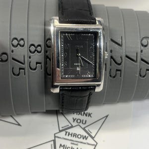 May include: A rectangular Timex watch with a black dial and silver-tone case. The watch has a black leather band. The watch is resting on a gray ring sizer with measurements in inches.
