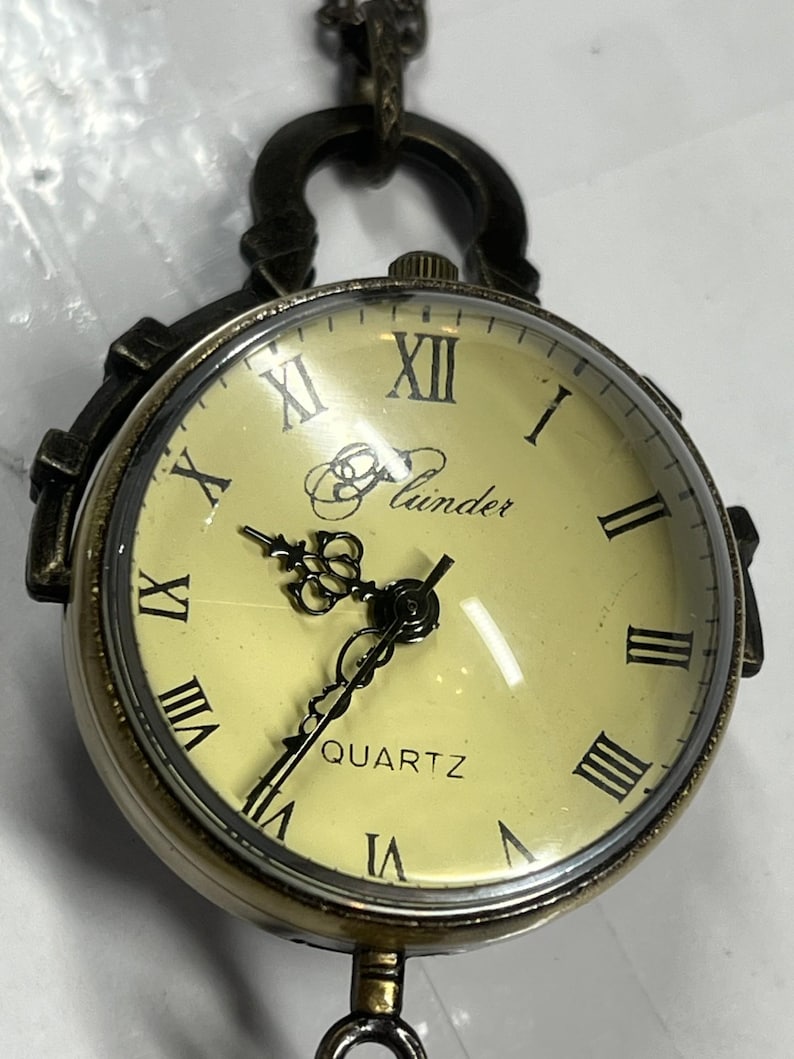 May include: A vintage-style pocket watch with a cream-colored face and black Roman numerals. The watch has a bronze-colored metal casing and a chain. The face reads "Quartz" and "Lunder".