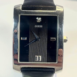 May include: A rectangular Guess watch with a black face and silver-tone case. The watch features a diamond accent and a black leather band. The watch face has vertical lines and the word "GUESS" is printed on the face.