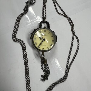 May include: Antique-style pocket watch pendant with a cream-colored face, Roman numerals, and the word "QUARTZ." The watch is suspended from a long, dark chain with a clasp and a decorative tassel. The watch case is a dark metal.