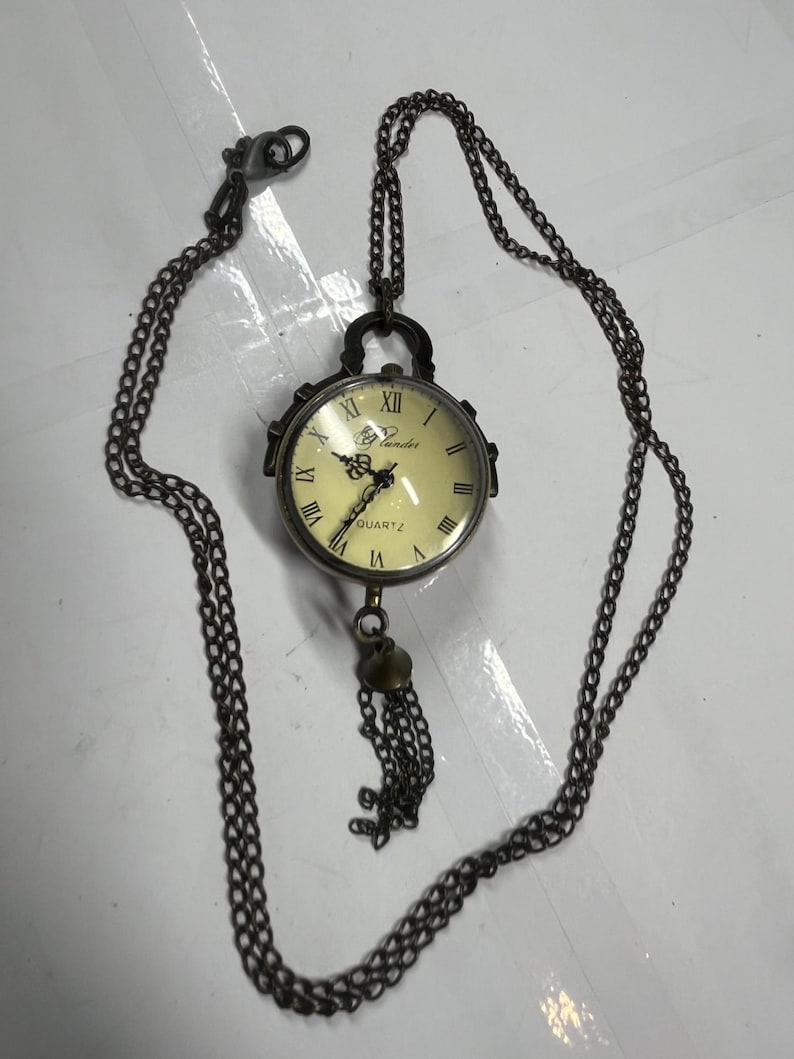 May include: Antique-style pocket watch pendant with a cream-colored face, Roman numerals, and the word "QUARTZ." The watch is suspended from a long, dark chain with a clasp and a decorative tassel. The watch case is a dark metal.