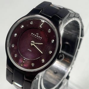 May include: A black Skagen Denmark watch with a dark purple face and silver hands. The watch face has small, sparkling accents and the word "STEEL" is printed on the face. The watch band is black and the watch is displayed on a clear stand.