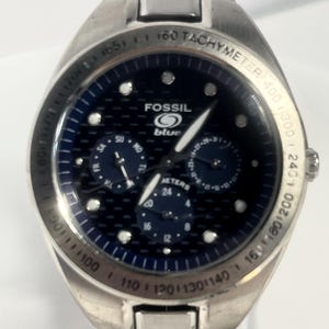 May include: A stainless steel Fossil Blue wristwatch with a dark blue face. The watch features three sub-dials, luminous markers, and a tachymeter bezel. The watch has a stainless steel band and a date display.