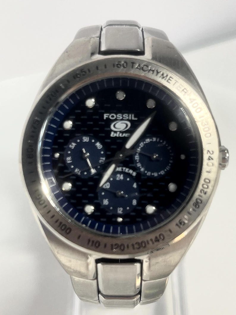 May include: A stainless steel Fossil Blue wristwatch with a dark blue face. The watch features three sub-dials, luminous markers, and a tachymeter bezel. The watch has a stainless steel band and a date display.