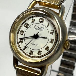 May include: A gold-toned Gitano quartz watch with a white face and black numerals. The watch features a gold bezel and a gold-colored, expandable band. The watch face displays the word "Gitano" and "Quartz".