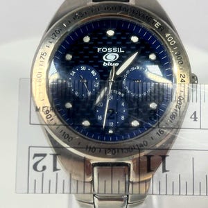 May include: A stainless steel Fossil Blue watch with a blue face. The watch has a tachymeter bezel and multiple subdials. The watch face displays the Fossil logo and the word "blue". A ruler is placed across the watch face.