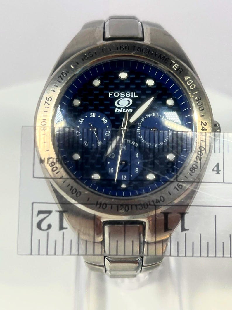 May include: A stainless steel Fossil Blue watch with a blue face. The watch has a tachymeter bezel and multiple subdials. The watch face displays the Fossil logo and the word "blue". A ruler is placed across the watch face.