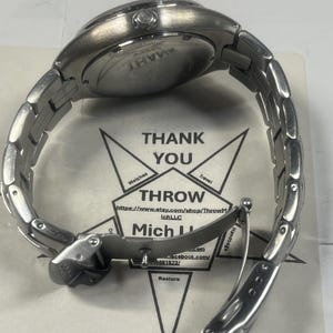 May include: A silver-toned wristwatch with a stainless steel band is shown from the back. The watch face is visible, and the band is partially unclasped. The watch rests on a paper with the words "THANK YOU" and "THROW" printed on it.