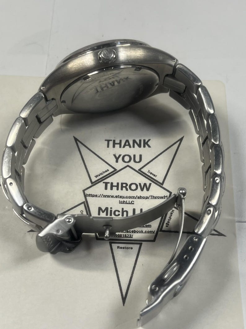 May include: A silver-toned wristwatch with a stainless steel band is shown from the back. The watch face is visible, and the band is partially unclasped. The watch rests on a paper with the words "THANK YOU" and "THROW" printed on it.