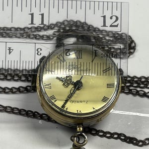 May include: A vintage-style quartz pendant watch with a cream-colored face, Roman numerals, and a decorative crown. The watch is suspended from a dark chain and has a small, decorative charm. The watch face is approximately 1 inch in diameter.