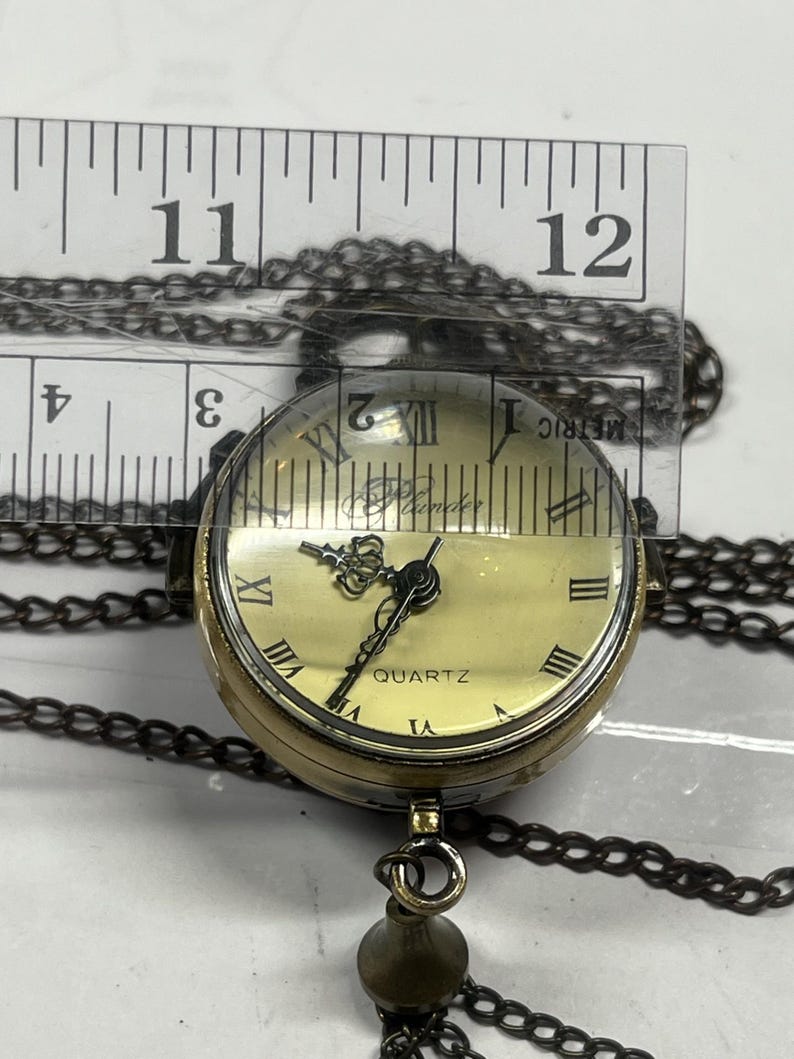 May include: A vintage-style quartz pendant watch with a cream-colored face, Roman numerals, and a decorative crown. The watch is suspended from a dark chain and has a small, decorative charm. The watch face is approximately 1 inch in diameter.