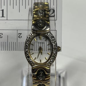 Może przedstawiać: Złoty zegarek Caravelle by Bulova z białą tarczą i diamentowymi akcentami. Zegarek ma złotą bransoletę i małą owalną tarczę. Zegarek ma szerokość około 2,5 cm.