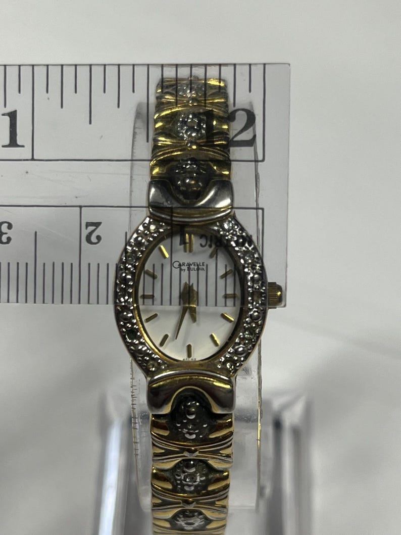 Może przedstawiać: Złoty zegarek Caravelle by Bulova z białą tarczą i diamentowymi akcentami. Zegarek ma złotą bransoletę i małą owalną tarczę. Zegarek ma szerokość około 2,5 cm.