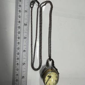 May include: A vintage-style pocket watch pendant necklace. The watch has a cream-colored face with Roman numerals and a quartz movement. The watch is suspended from a dark chain. The watch measures approximately 1 inch in diameter.