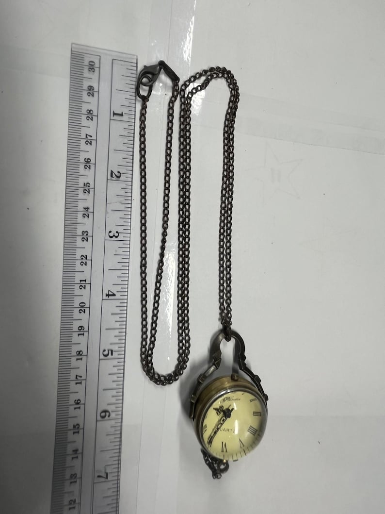 May include: A vintage-style pocket watch pendant necklace. The watch has a cream-colored face with Roman numerals and a quartz movement. The watch is suspended from a dark chain. The watch measures approximately 1 inch in diameter.