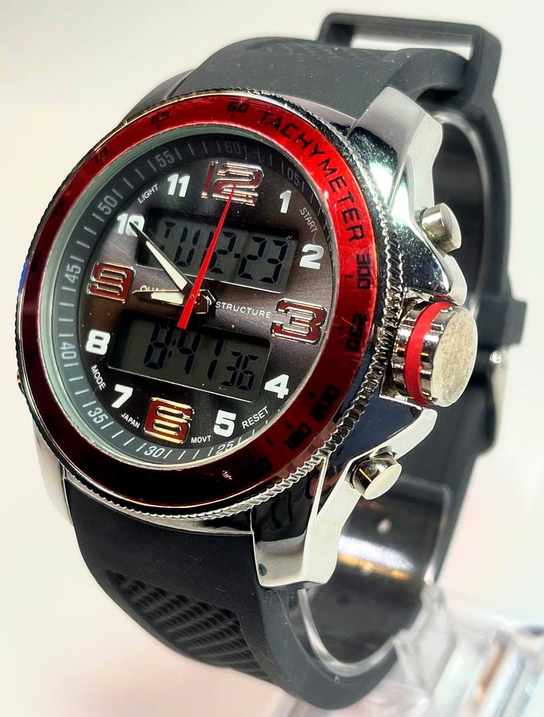 Peut inclure: Montre num&eacute;rique noire et argent&eacute;e avec une lunette rouge et un bracelet en caoutchouc noir. Le cadran affiche l'heure et porte l'inscription "TACHYMETER". La montre poss&egrave;de plusieurs boutons et un affichage num&eacute;rique.