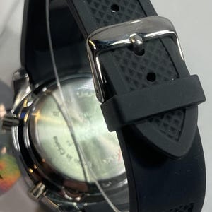Peut inclure: Gros plan d'une montre-bracelet noire avec un bracelet en caoutchouc textur&eacute; et une boucle argent&eacute;e. Le cadran de la montre est visible, avec un bo&icirc;tier argent&eacute; et un cadran noir. Le bracelet est fix&eacute; avec un fermoir en m&eacute;tal.