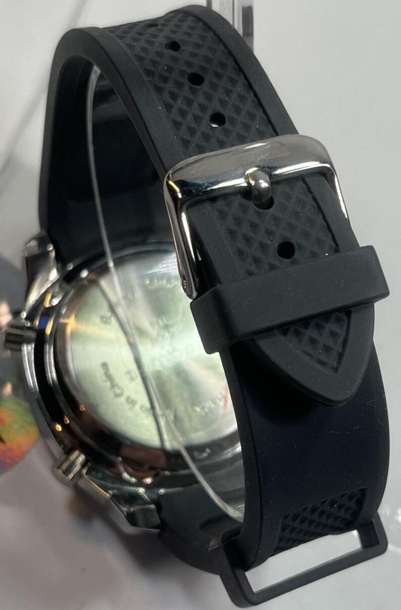 Peut inclure: Gros plan d'une montre-bracelet noire avec un bracelet en caoutchouc textur&eacute; et une boucle argent&eacute;e. Le cadran de la montre est visible, avec un bo&icirc;tier argent&eacute; et un cadran noir. Le bracelet est fix&eacute; avec un fermoir en m&eacute;tal.