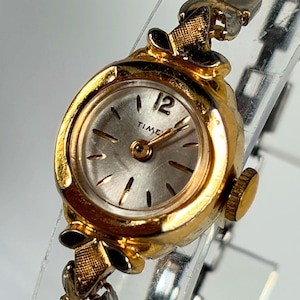 May include: A gold-tone analog wristwatch with a round case and a silver dial featuring the word TIME and the number 12. The watch has a textured gold-tone bezel and a flexible metal link bracelet with a metallic finish and gold-tone accents.