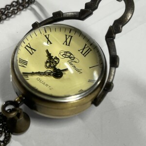May include: A vintage-style pocket watch with a cream-colored face and Roman numerals. The watch has a brass-colored casing and a dark chain. The watch face reads "Quartz" and the brand name "Plunder".
