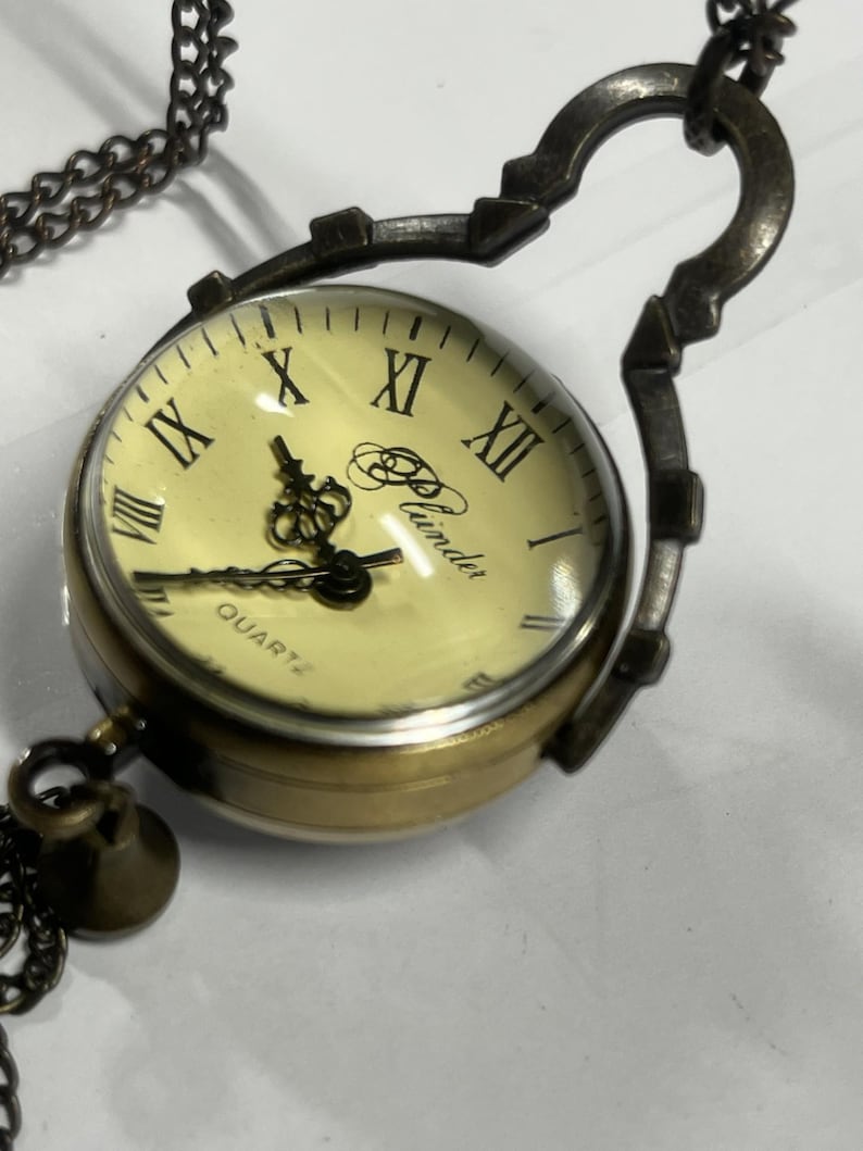 May include: A vintage-style pocket watch with a cream-colored face and Roman numerals. The watch has a brass-colored casing and a dark chain. The watch face reads "Quartz" and the brand name "Plunder".