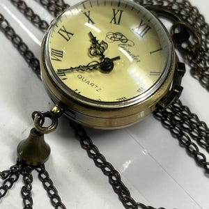May include: A vintage-style quartz pocket watch pendant with a bronze finish. The watch features a cream-colored face with black Roman numerals and hands. The watch is suspended from a long, bronze-colored chain with a decorative tassel.