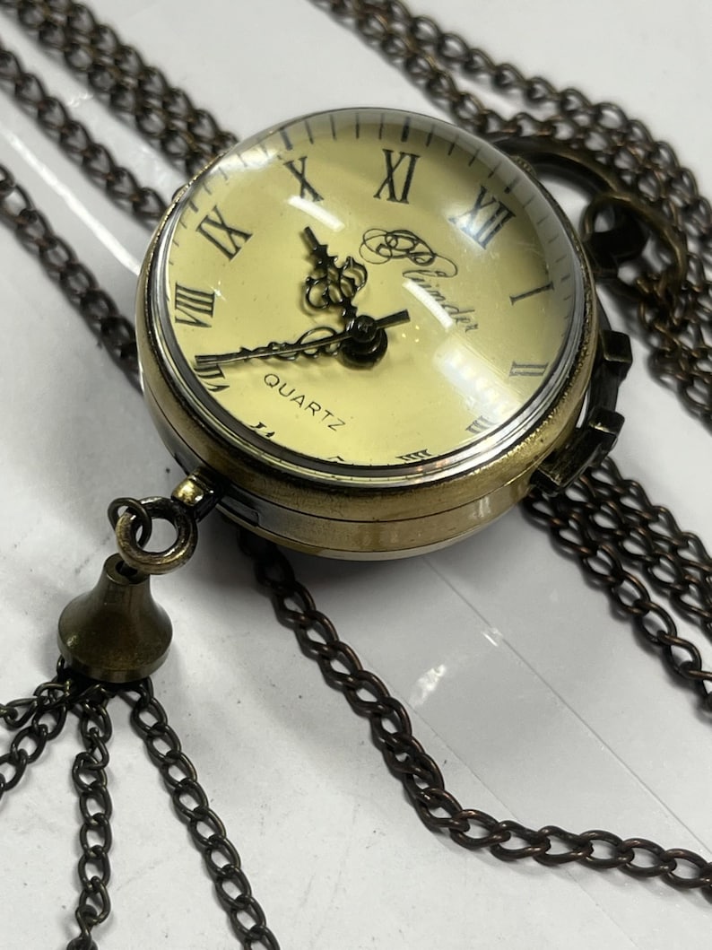 May include: A vintage-style quartz pocket watch pendant with a bronze finish. The watch features a cream-colored face with black Roman numerals and hands. The watch is suspended from a long, bronze-colored chain with a decorative tassel.