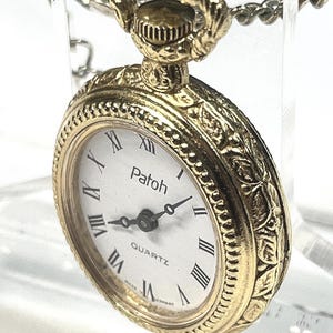 May include: A gold-toned Patoh quartz pocket watch with a white face and black Roman numerals. The watch has an ornate floral design around the bezel and a chain. The watch reads "QUARTZ" and "MADE IN GERMANY".