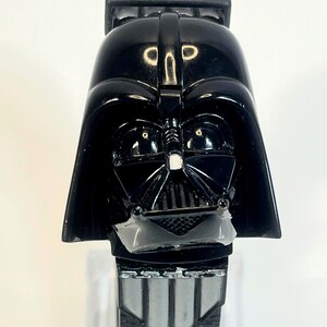 Pu&ograve; includere: Un orologio nero di Darth Vader con il design del casco e della maschera del personaggio. L'orologio ha un quadrante rettangolare con un cinturino argentato e nero. L'orologio &egrave; un oggetto da collezione del franchise di Star Wars.