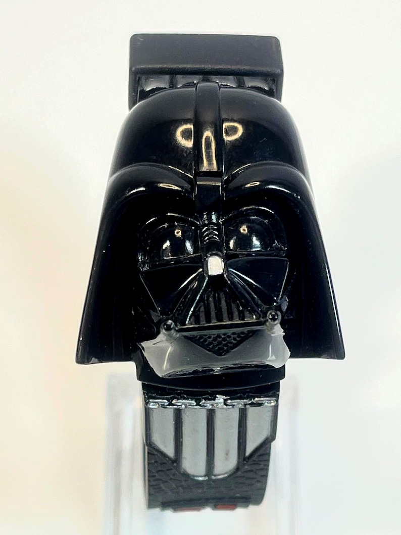 Pu&ograve; includere: Un orologio nero di Darth Vader con il design del casco e della maschera del personaggio. L'orologio ha un quadrante rettangolare con un cinturino argentato e nero. L'orologio &egrave; un oggetto da collezione del franchise di Star Wars.
