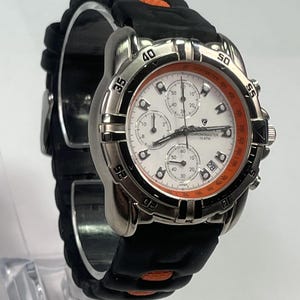 May include: A stainless steel Chronomaster watch with a white face and orange accents. The watch features multiple sub-dials and a date window. It has a black rubber strap with orange details. The watch is displayed on a clear stand.