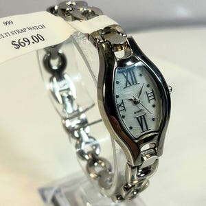 May include: Silver-toned quartz watch with a white oval face and Roman numeral hour markers. The watch features a linked bracelet-style band. The tag reads "MULTI STRAP WATCH".